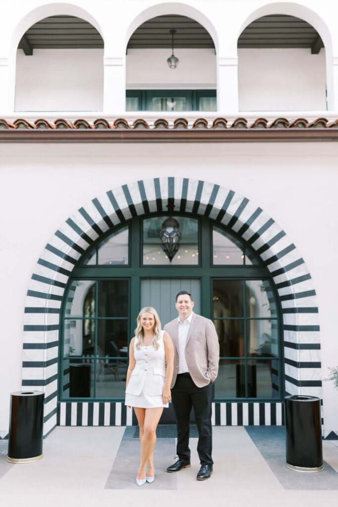 A woman in a white outfit and a man in a light blazer and black pants stand smiling in front of a large arched doorway with black-and-white stripes on a white building with arched balconies above. Copyright - Lisa Leanne Photography, California