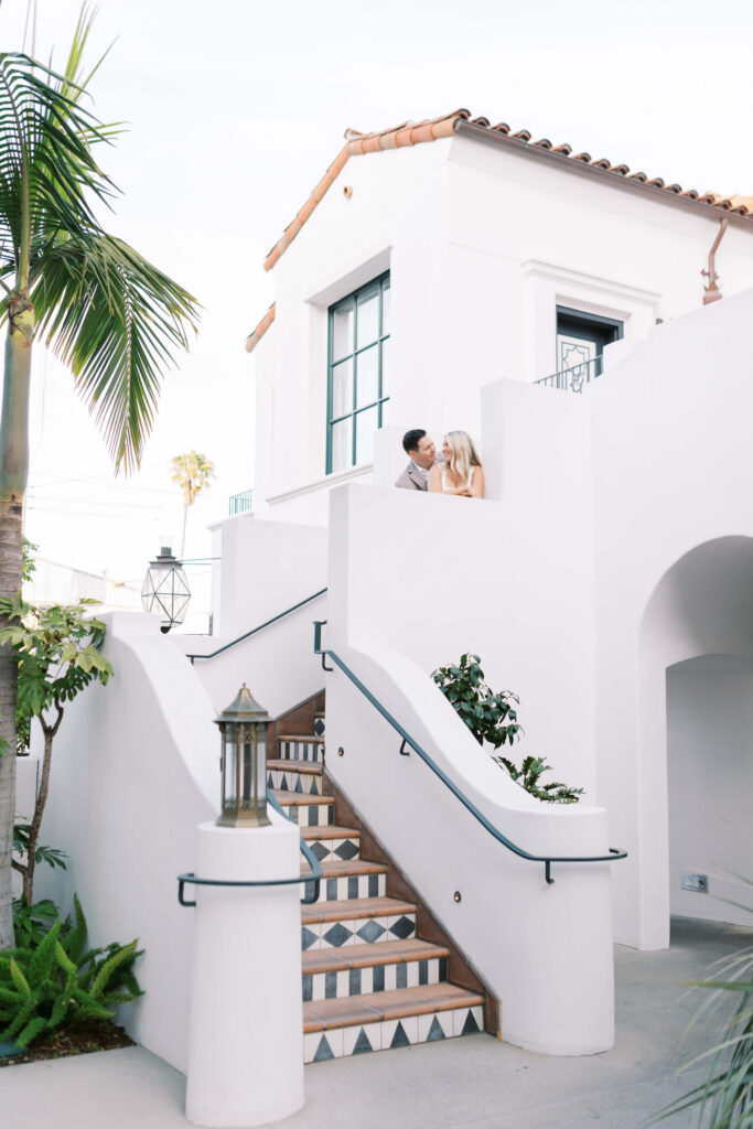 A couple stands on the balcony of a white, Mediterranean-style building with terracotta roof tiles. A staircase with patterned tiles and a lantern leads up, surrounded by tropical plants and palm trees. Copyright - Lisa Leanne Photography, California