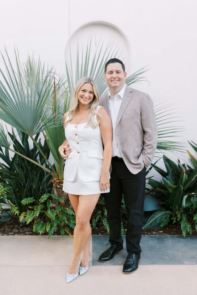 A smiling woman in a white sleeveless dress stands next to a man in a light blazer and black pants. They pose together outdoors in front of green plants and a white wall. Copyright - Lisa Leanne Photography, California