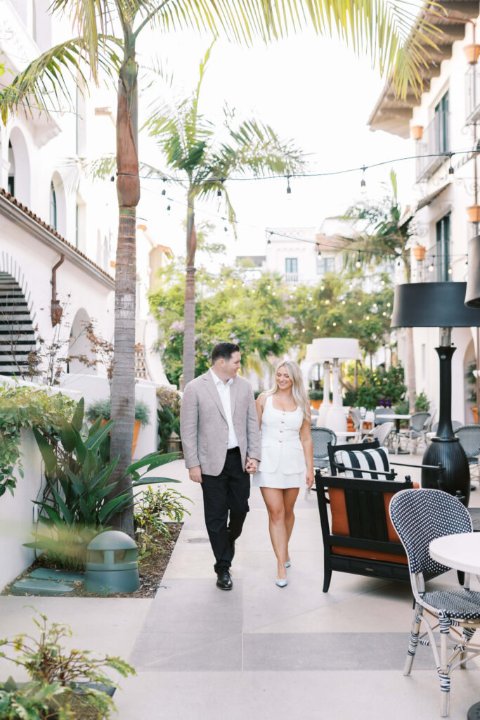 A couple walks hand in hand down a stylish outdoor courtyard lined with palm trees, greenery, and elegant furniture. The man wears a light blazer and black pants, while the woman wears a white dress and heels. Copyright - Lisa Leanne Photography, California