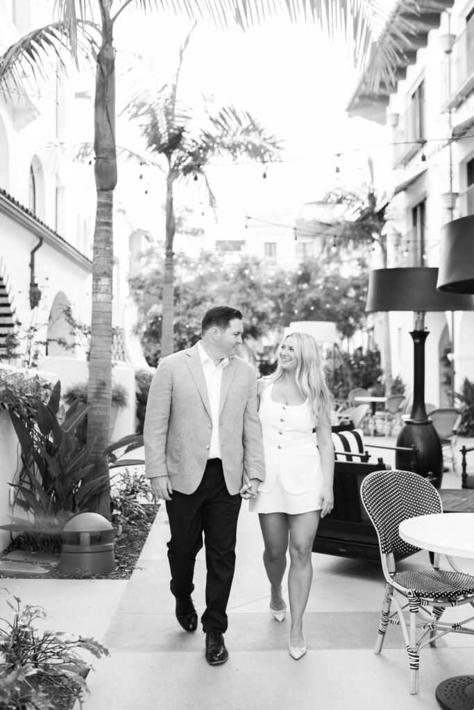 A man and woman walk hand in hand outdoors along a stylish, palm-lined path. Both are smiling and dressed in light-colored, semi-formal outfits. The scene has a relaxed, elegant atmosphere. Copyright - Lisa Leanne Photography, California