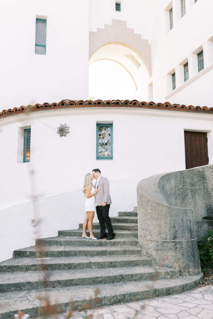 A couple stands close together, touching foreheads, on a curved outdoor staircase beside a white building with blue-trimmed windows and a wooden door. The setting appears romantic and peaceful. Copyright - Lisa Leanne Photography, California