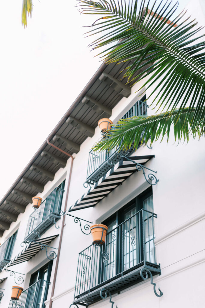 A white building with black iron balconies, striped awnings, and potted plants, framed by green palm leaves against a bright sky. Copyright - Lisa Leanne Photography, California