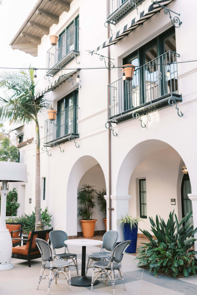 A stylish outdoor patio with potted plants, a round table with chairs, palm trees, and a white building with arched doorways, balconies, and green window frames in the background. Copyright - Lisa Leanne Photography, California