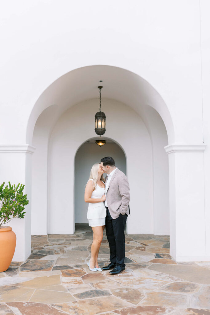A couple stands close together under a white archway, with the woman in a white dress and the man in a blazer and dark pants, sharing an intimate moment. A potted plant sits to the left of the arch. Copyright - Lisa Leanne Photography, California