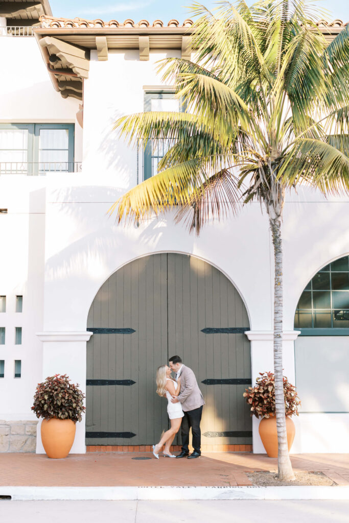 A couple shares a kiss in front of large arched double doors on a white building, flanked by potted plants, with a tall palm tree casting shadows across the scene. Copyright - Lisa Leanne Photography, California
