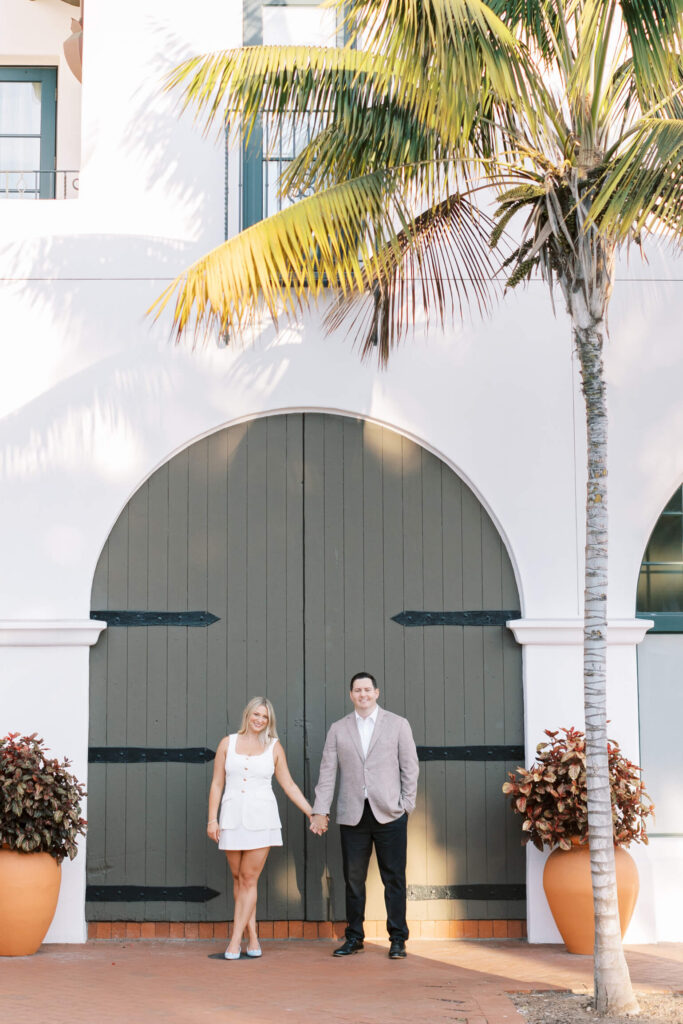 A smiling couple holds hands in front of a large arched wooden door, with a palm tree and potted plants nearby. Copyright - Lisa Leanne Photography, California