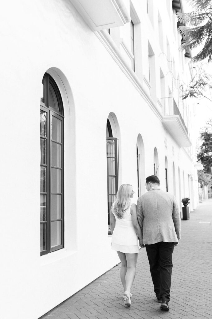 A couple walks hand-in-hand down a sidewalk beside a white building with tall arched windows. The image is in black and white, and they are seen from behind. Copyright - Lisa Leanne Photography, California