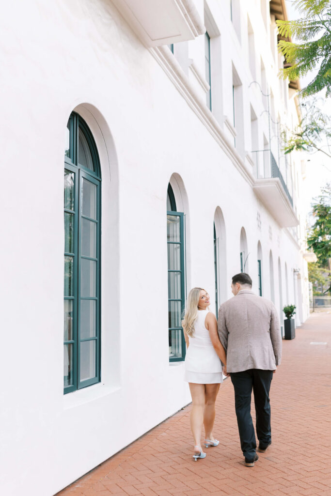 A couple walks hand in hand on a brick sidewalk beside a white building with arched windows. The woman wears a white dress and heels, and the man wears a beige blazer and black pants. Copyright - Lisa Leanne Photography, California