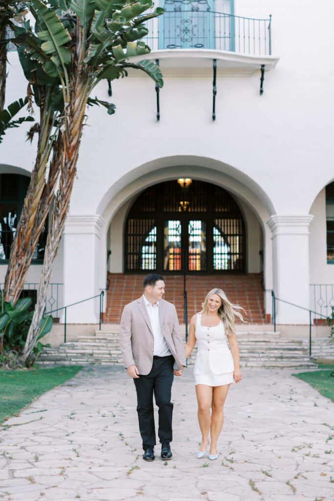 A couple holding hands and smiling, walking on a stone path in front of a large white building with arches, palms, and a balcony. The man wears a blazer and pants, and the woman wears a white dress. Copyright - Lisa Leanne Photography, California