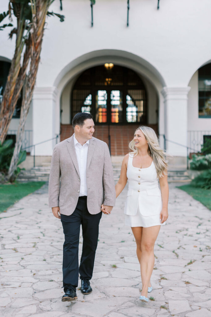 A man and woman holding hands and smiling at each other as they walk on a stone path in front of a white building with arches. Copyright - Lisa Leanne Photography, California