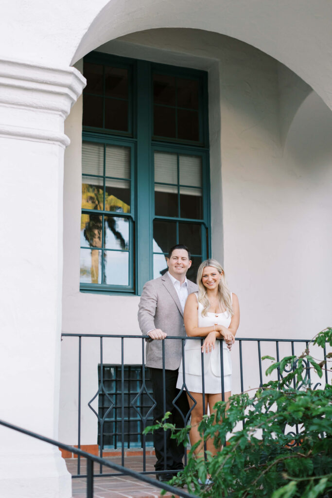 A man in a light-colored blazer and a woman in a white dress stand together, smiling, on a balcony with an iron railing in front of a large window and white building exterior. Green foliage is visible below them. Copyright - Lisa Leanne Photography, California