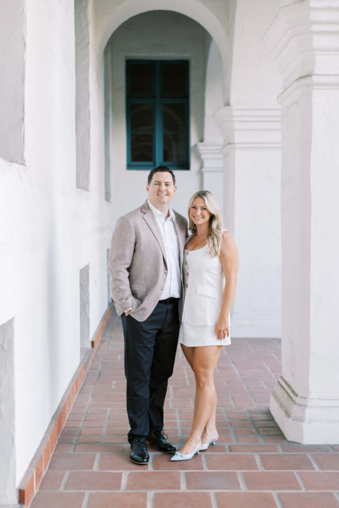 A man in a light blazer and dark pants stands next to a woman in a white dress, both smiling, in a bright, arched hallway with white walls and terra-cotta tile flooring. Copyright - Lisa Leanne Photography, California