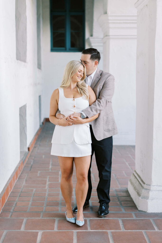 A couple stands in a sunlit hallway with white walls and tiled floor. The woman in a white dress smiles while the man, in a blazer and black pants, embraces her from behind and kisses her head. Copyright - Lisa Leanne Photography, California