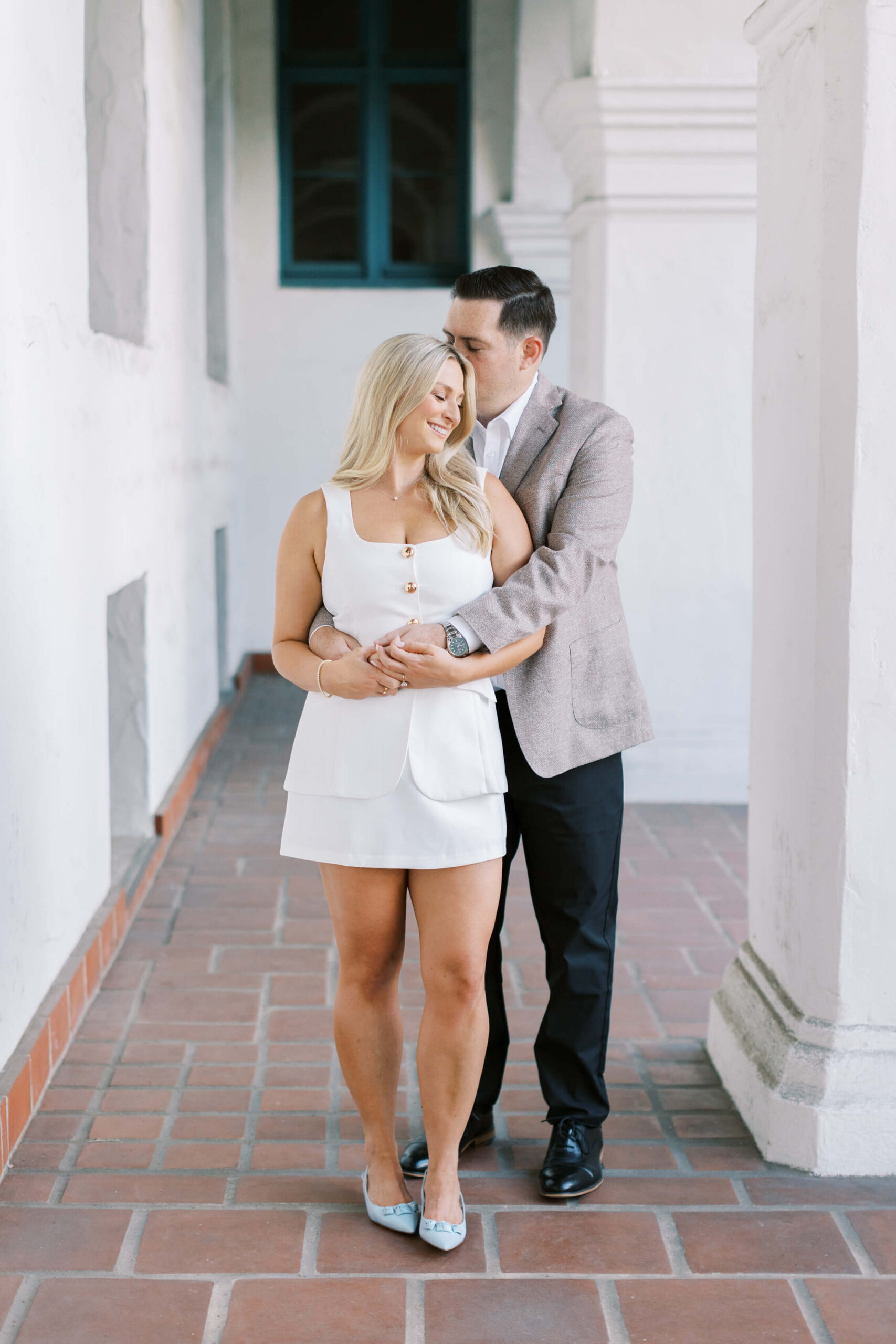 A couple stands in a sunlit hallway with white walls and tiled floor. The woman in a white dress smiles while the man, in a blazer and black pants, embraces her from behind and kisses her head. Copyright - Lisa Leanne Photography, California