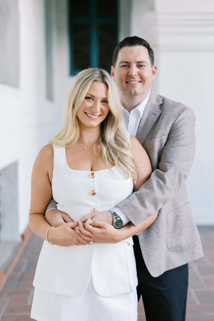 A smiling couple poses together. The woman, with long blonde hair in a white dress, stands in front while the man, in a grey blazer, stands behind her, wrapping his arms around her waist. They are indoors with a white background. Copyright - Lisa Leanne Photography, California