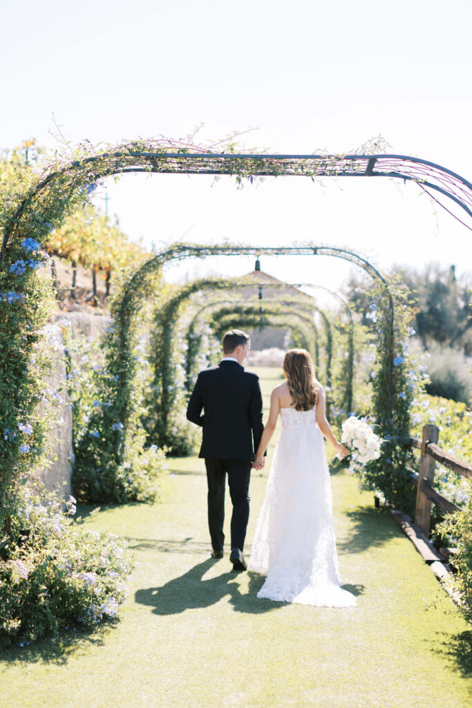A bride and groom walk hand in hand down a sunlit garden path under green archways at their Cielo Farms Wedding, surrounded by flowers and greenery. Captured beautifully by Lisa Leanne Photography, the bride glows in white beside her groom in black. Copyright - Lisa Leanne Photography, California