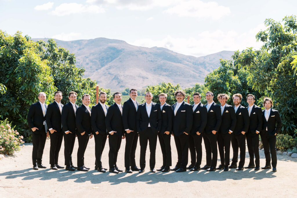 Gerry Ranch Wedding - A group of fifteen Groomsmen in black suits with white shirts and bow ties stand in a line outdoors, smiling, with green trees and mountains visible in the background under a blue sky. Copyright - Lisa Leanne Photography, California