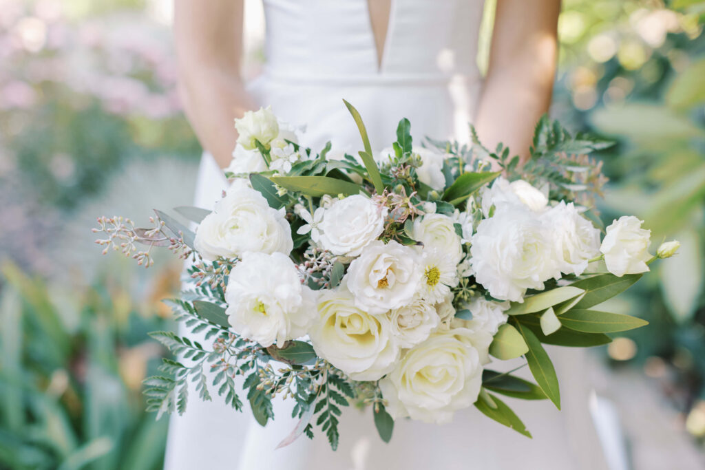 Gerry Ranch Wedding - A person in a white dress holds a bouquet of white roses and greenery, standing outdoors with blurred plants and flowers in the background. Copyright - Lisa Leanne Photography, California
