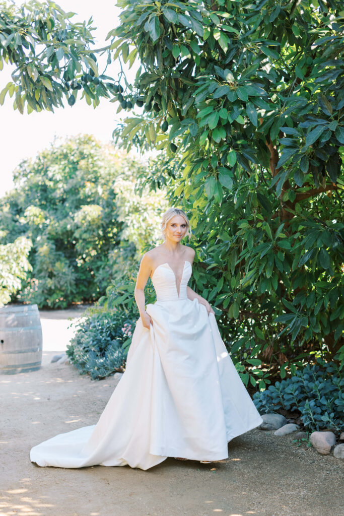 Gerry Ranch Wedding - A Bride in a strapless white wedding gown stands outdoors on a dirt path, holding her dress with both hands. She is surrounded by lush green trees and sunlight filters through the leaves. Copyright - Lisa Leanne Photography, California