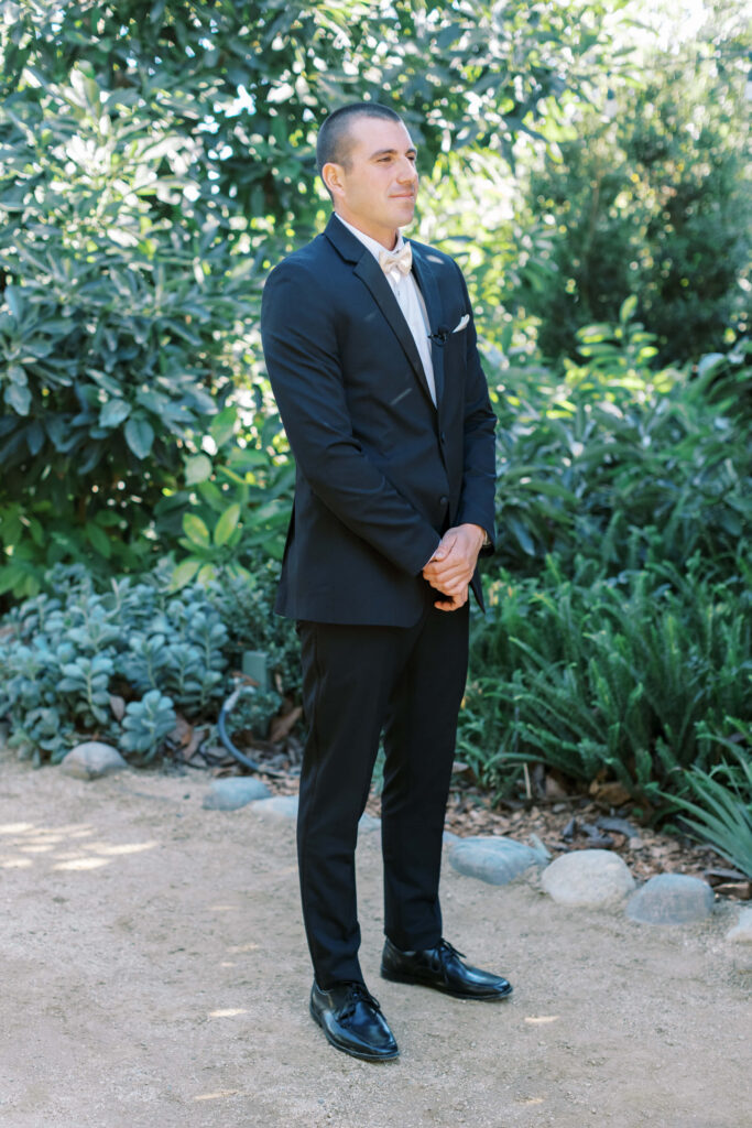 Gerry Ranch Wedding - A Groom in a dark suit stands on a sandy path outdoors, hands clasped, surrounded by lush green plants and trees, with sunlight filtering through the foliage. Copyright - Lisa Leanne Photography, California