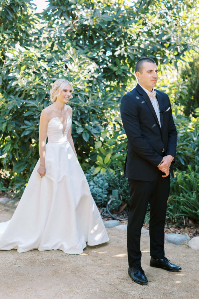 Gerry Ranch Wedding First Look- A bride in a white strapless gown approaches a groom in a black suit, who stands with his back to her, outdoors surrounded by lush greenery. Copyright - Lisa Leanne Photography, California