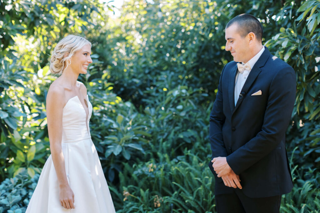 Gerry Ranch Wedding First Look - A bride in a white dress and a groom in a black suit with a bow tie smile at each other outdoors, surrounded by lush green foliage. Copyright - Lisa Leanne Photography, California