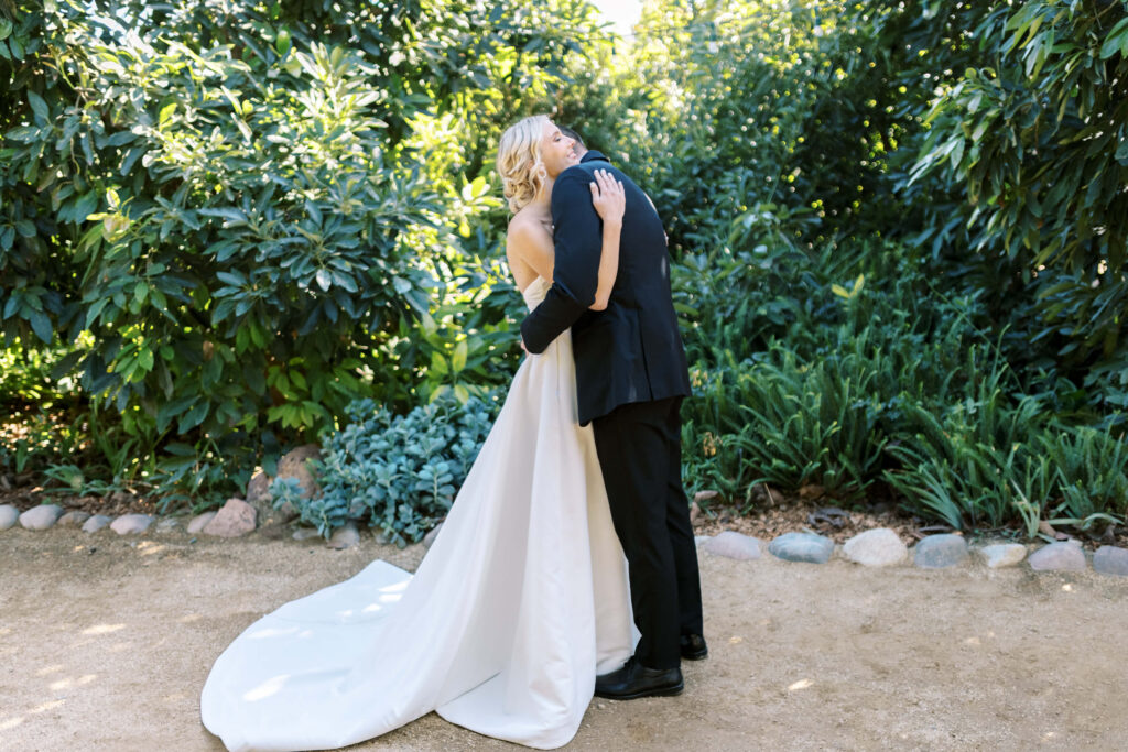 Gerry Ranch Wedding First Look - A bride in a white gown and a groom in a black suit embrace outdoors on a sunny day, surrounded by lush green foliage and a stone-bordered path. Copyright - Lisa Leanne Photography, California