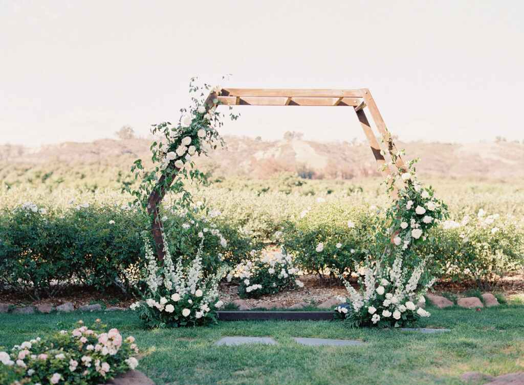 Gerry Ranch Wedding Ceremony - A wooden hexagon wedding arch decorated with white flowers and greenery stands on a grassy lawn with bushes and distant hills in the background. Copyright - Lisa Leanne Photography, California