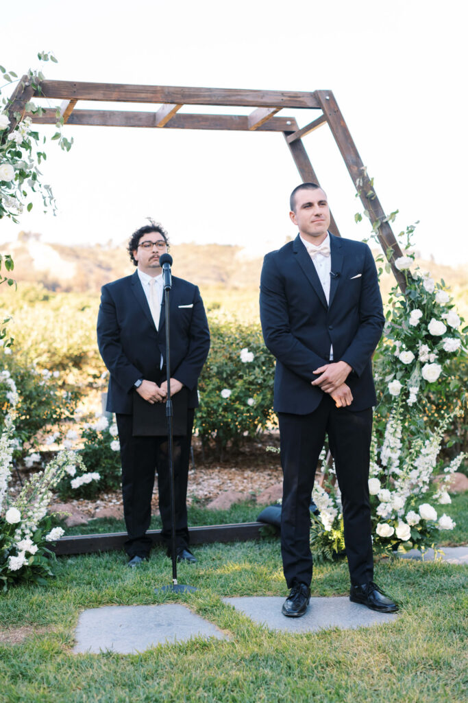 Gerry Ranch Wedding Ceremony -Two men in suits stand under a wooden wedding arch decorated with white flowers and greenery outdoors. One stands at a microphone, the other stands slightly in front, both looking forward. Copyright - Lisa Leanne Photography, California