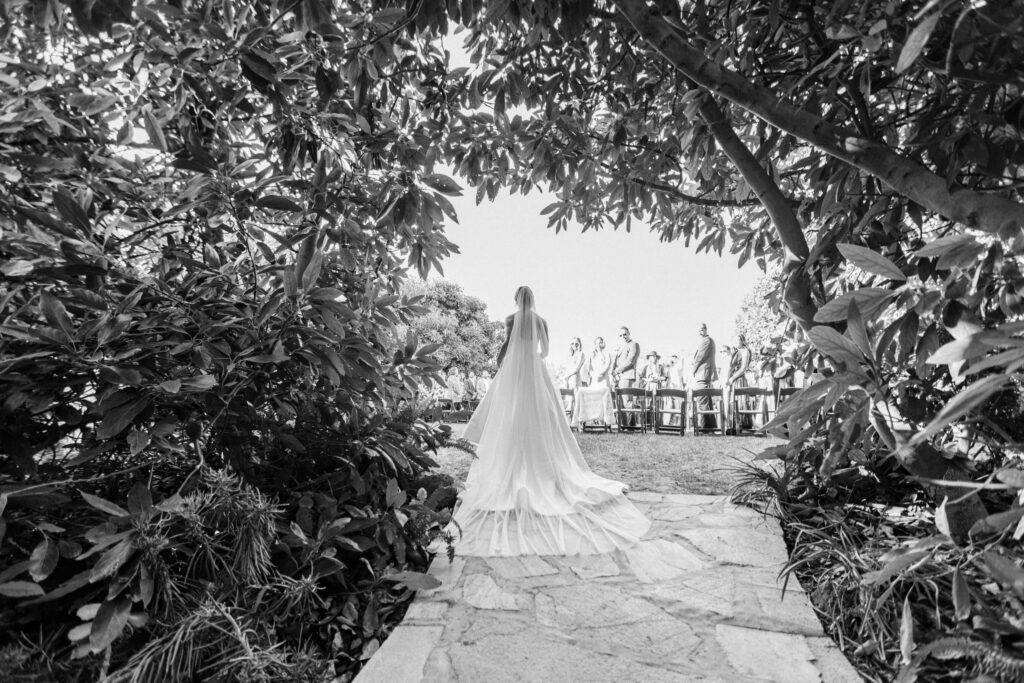 Gerry Ranch Wedding Ceremony -A bride in a long, flowing gown walks down an outdoor stone path toward guests seated for a wedding ceremony, framed by leafy trees. The scene is in black and white. Copyright - Lisa Leanne Photography, California