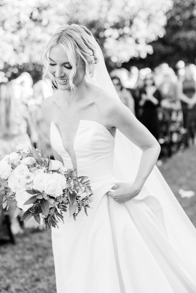 Gerry Ranch Wedding Ceremony -A bride in a strapless wedding gown holds a bouquet of flowers and smiles, walking outdoors. People and trees are blurred in the background. The photo is in black and white. Copyright - Lisa Leanne Photography, California