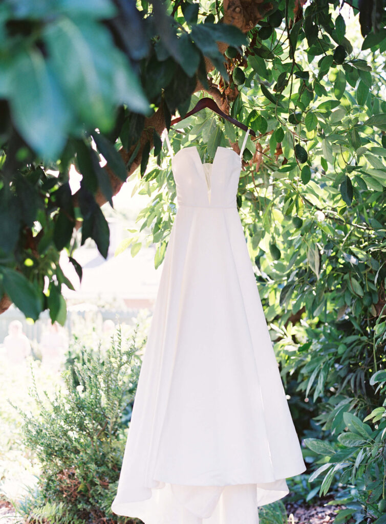 Gerry Ranch Wedding - A white sleeveless wedding dress with a deep V neckline hangs from a tree branch, surrounded by lush green foliage in an outdoor garden setting. Copyright - Lisa Leanne Photography, California