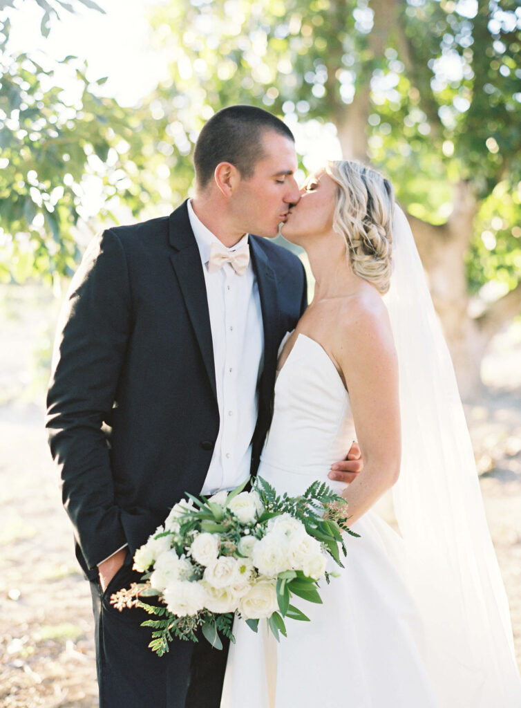Gerry Ranch Bride and Groom Portrait - A bride and groom share a kiss outdoors. The bride wears a strapless white gown, veil, and holds a bouquet of white flowers. The groom wears a black suit with a white shirt and bow tie. Sunlight filters through trees in the background. Copyright - Lisa Leanne Photography, California