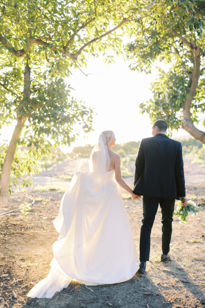 Gerry Ranch Bride and Groom Portrait - A bride in a white dress and veil holds hands with a groom in a dark suit as they walk outdoors under trees in sunlight, surrounded by nature. Copyright - Lisa Leanne Photography, California