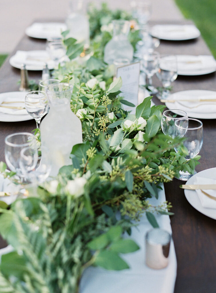Gerry Ranch Reception - A long wooden table set for a meal with white plates, glasses, and napkins. A lush green and white floral garland runs down the center, along with pitchers of water and candles. The setting is outdoors. Copyright - Lisa Leanne Photography, California
