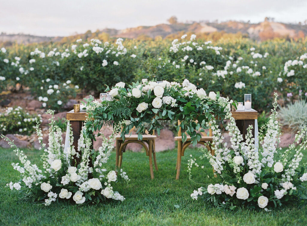 Gerry Ranch Reception - Bride and Groom table beautifully decorated with white roses and greenery, surrounded by lush floral arrangements and set in a garden with blooming white flowers and green hills in the background. Copyright - Lisa Leanne Photography, California