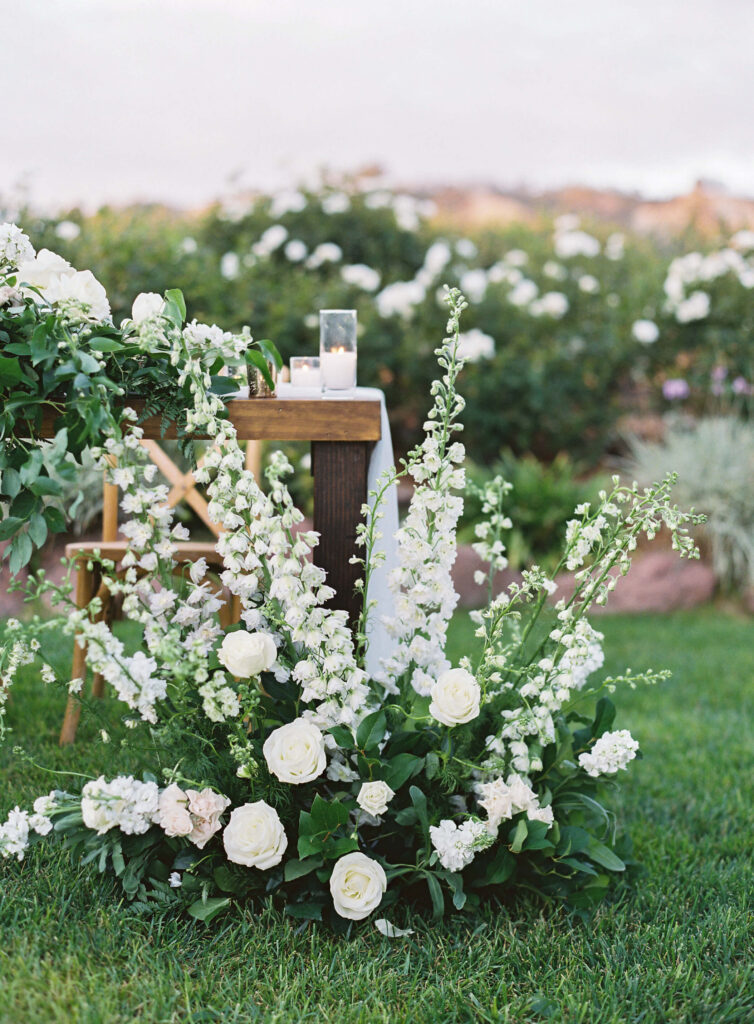 Gerry Ranch Reception - An outdoor floral arrangement features tall white flowers and roses with lush greenery, placed beside a wooden table with candles, set on a green lawn with more flowers in the background. Copyright - Lisa Leanne Photography, California