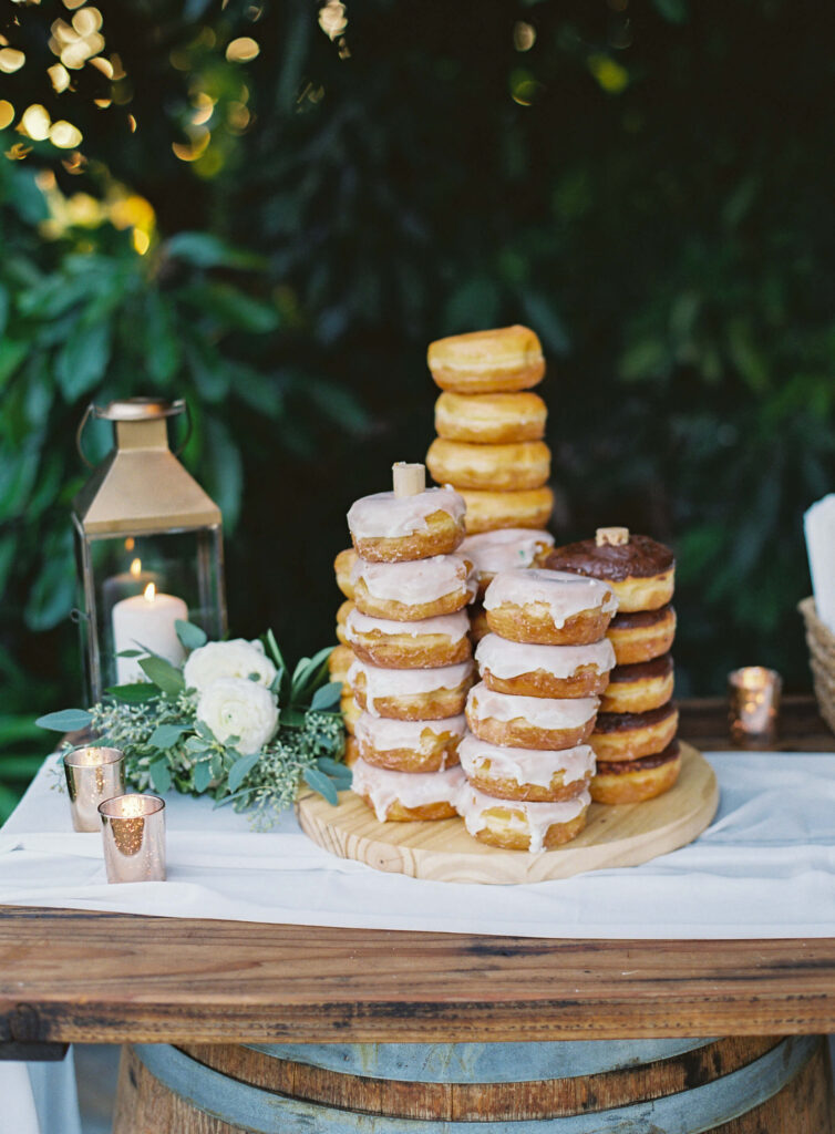 Gerry Ranch Reception - A rustic dessert table displays stacks of glazed donuts on a wooden board, surrounded by a lantern with a lit candle, white flowers, greenery, and small votive candles, set outdoors against lush green foliage. Copyright - Lisa Leanne Photography, California