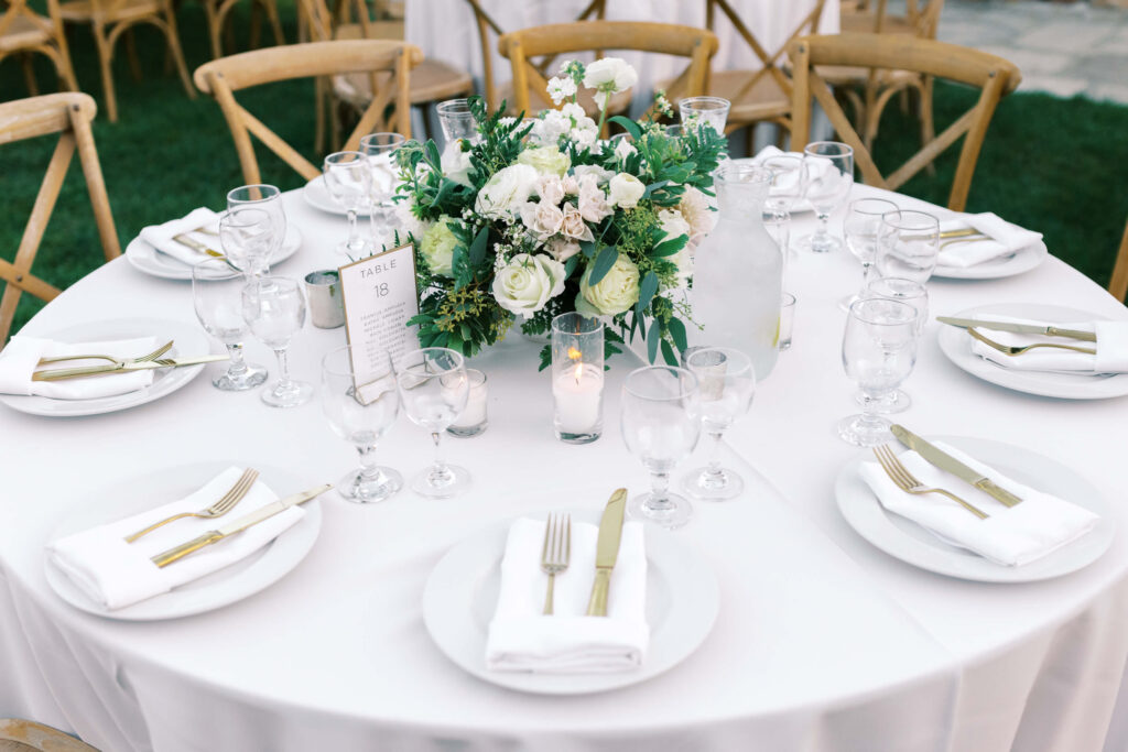 Gerry Ranch Reception - A round table set for eight with white plates, gold cutlery, napkins, and glassware. A floral centerpiece with white flowers and greenery is in the middle, surrounded by candles. Wooden chairs encircle the table. Copyright - Lisa Leanne Photography, California