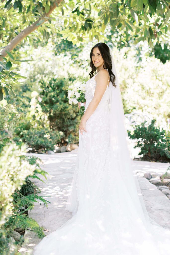 Gerry Ranch Bridal Portrait - A bride in a white wedding dress stands outdoors on a sunlit stone path, holding a bouquet of flowers, surrounded by lush green foliage and trees. Copyright - Lisa Leanne Photography, California