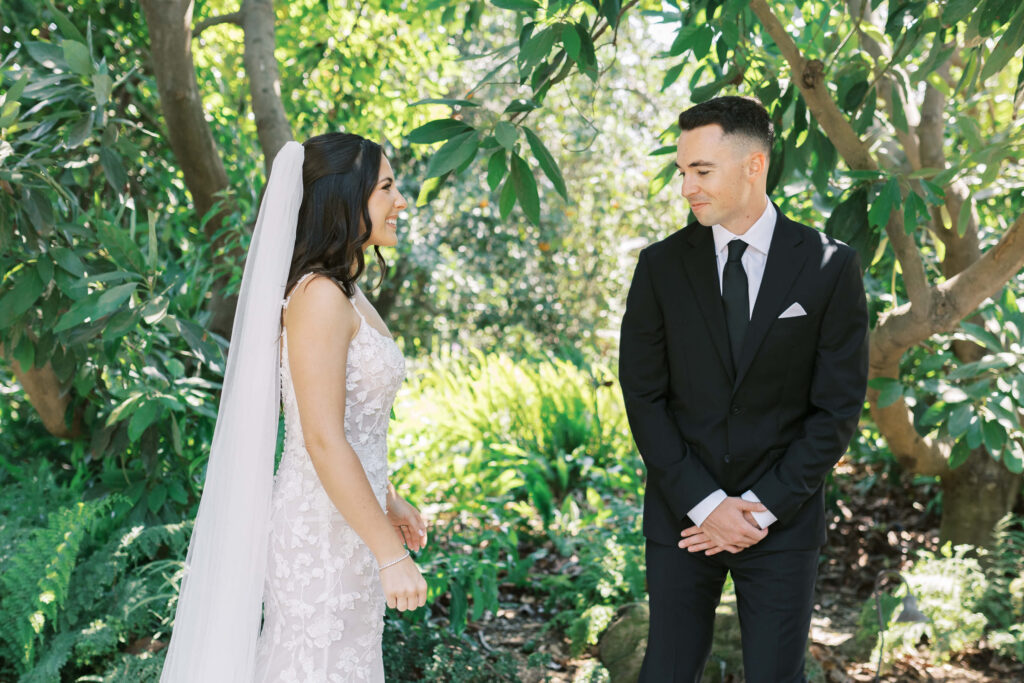 First Look at Gerry Ranch - A bride in a white dress and long veil stands smiling across from a groom in a black suit and tie. They are outdoors in a lush, green garden with sunlight filtering through the trees. Copyright - Lisa Leanne Photography, California