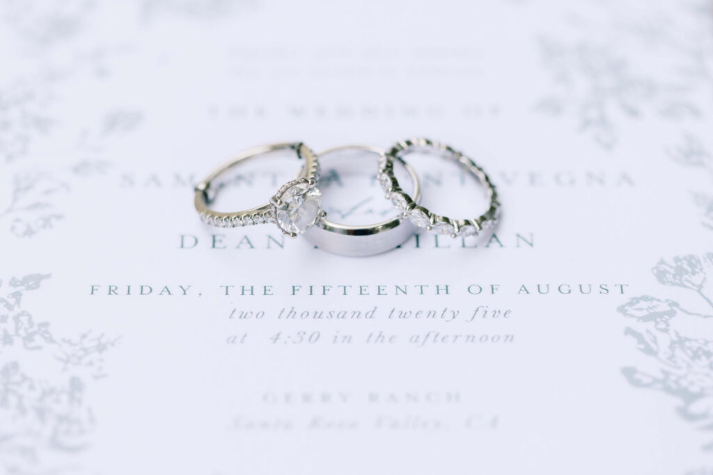 Gerry Ranch Wedding - Three wedding rings, including an engagement ring with a central diamond, are placed on a wedding invitation with elegant script and floral borders. The text and details are softly blurred in the background. Copyright - Lisa Leanne Photography, California