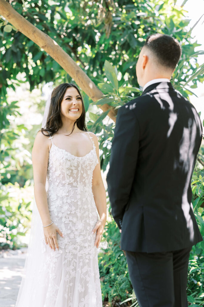 First Look at Gerry Ranch - A bride in a white floral lace dress smiles brightly at a groom in a black suit as they stand together outdoors, surrounded by lush green foliage and sunlight. Copyright - Lisa Leanne Photography, California