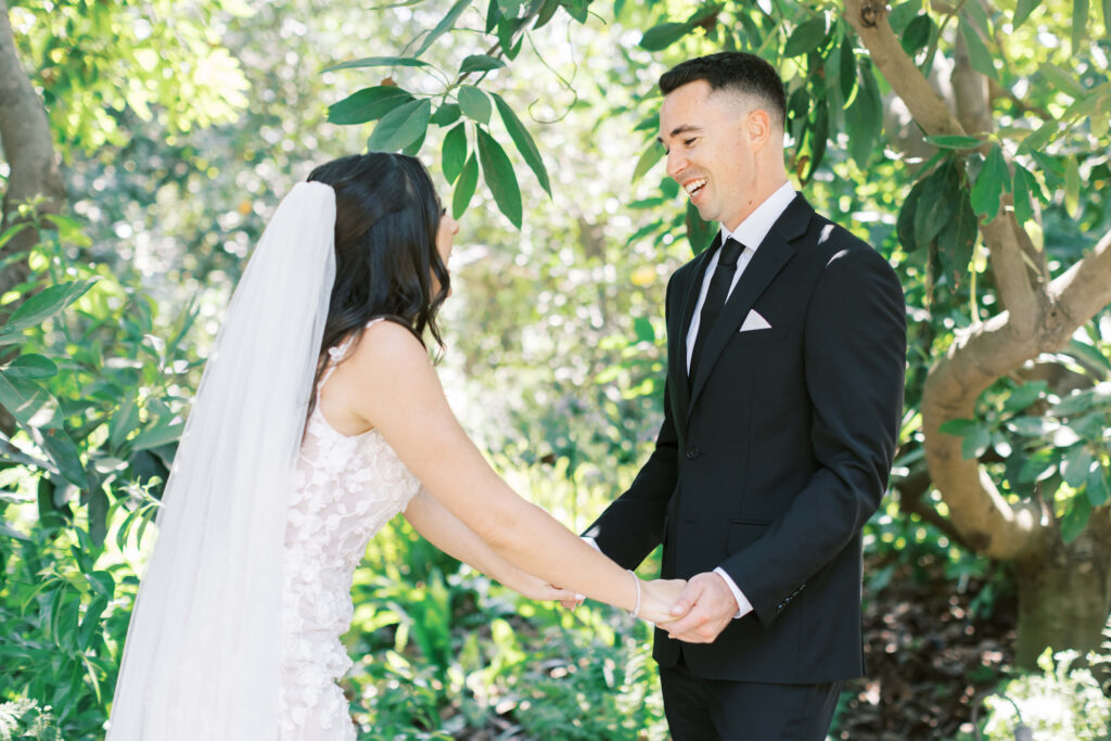 First Look at Gerry Ranch - A bride in a white dress and veil holds hands with a groom in a black suit and tie, both smiling and standing outdoors surrounded by lush green trees and sunlight. Copyright - Lisa Leanne Photography, California