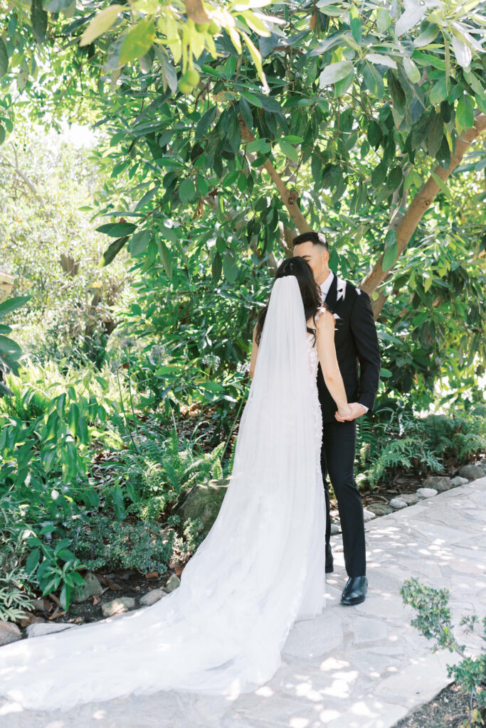 First Look at Gerry Ranch - A bride and groom stand on a sunlit garden path, holding hands and kissing. The bride wears a long white dress with a veil, and the groom is in a black suit. Greenery and trees surround them. Copyright - Lisa Leanne Photography, California