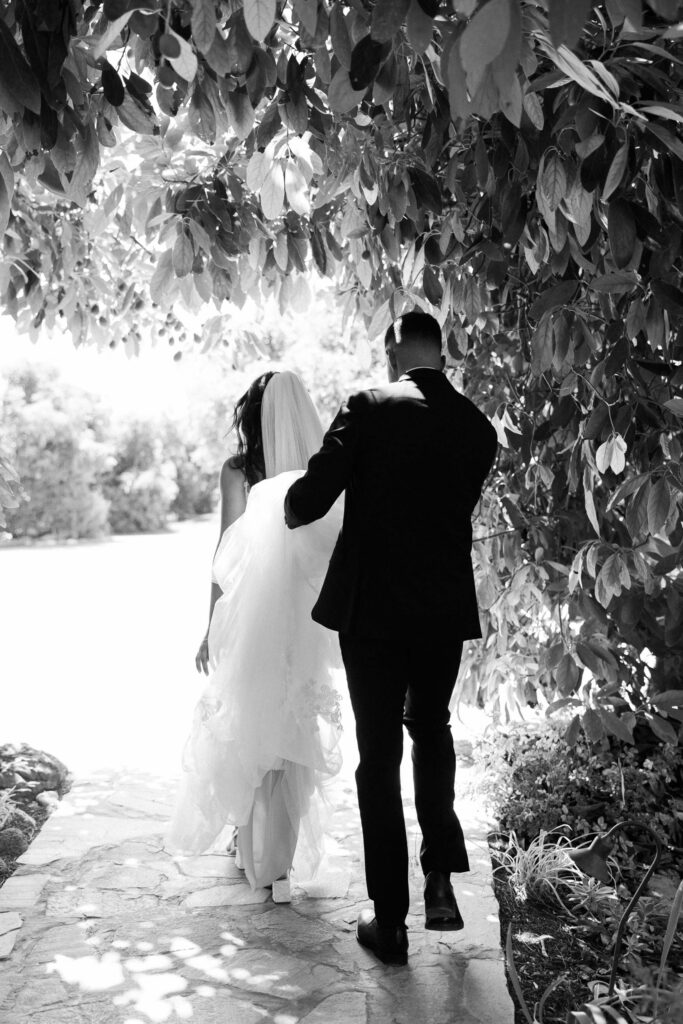 First Look at Gerry Ranch - A bride in a veil and a groom in a suit walk arm in arm along a stone path, surrounded by leafy trees, with sunlight shining ahead. Copyright - Lisa Leanne Photography, California