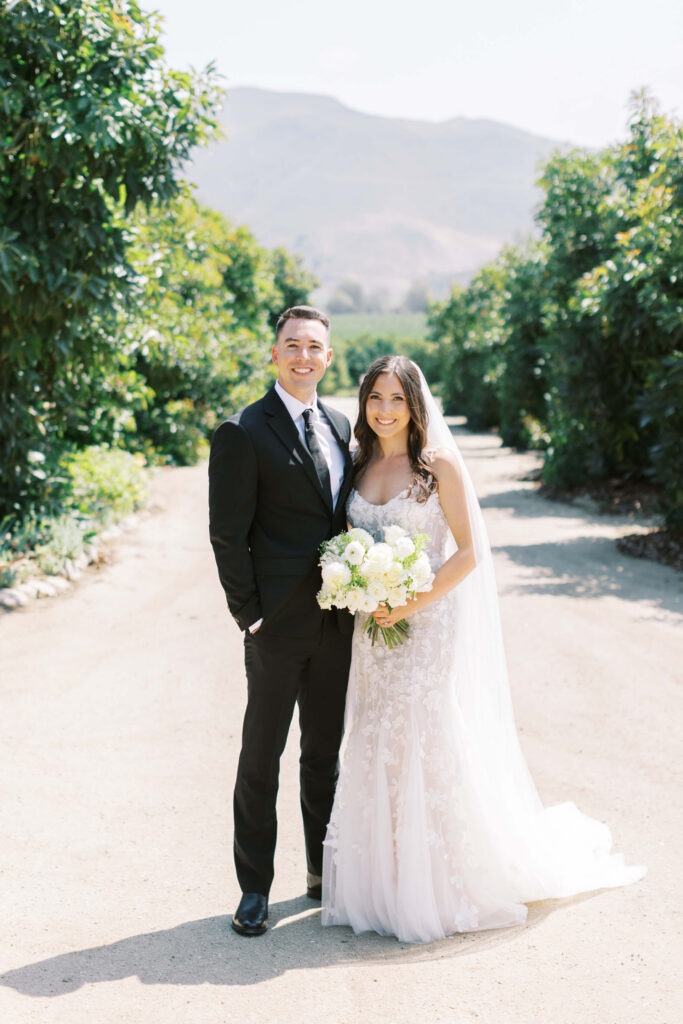 Bride and Groom Portrait at Gerry Ranch - A bride and groom stand together outdoors on a sunny day. The bride wears a white dress and holds a bouquet, while the groom wears a black suit. Green trees and mountains are visible in the background. Copyright - Lisa Leanne Photography, California
