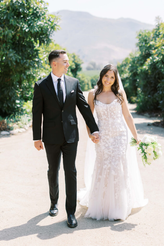 Bride and Groom Portrait at Gerry Ranch - A bride in a white lace dress and a groom in a black suit walk hand in hand outdoors on a sunny day, smiling at each other. Lush green trees and mountains are visible in the background. Copyright - Lisa Leanne Photography, California