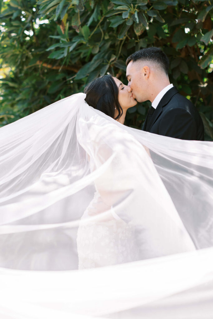 Bride and Groom Portrait at Gerry Ranch - A bride and groom share a kiss outdoors, surrounded by greenery. The bride’s long veil billows around them, creating a soft, romantic frame for their embrace. Copyright - Lisa Leanne Photography, California